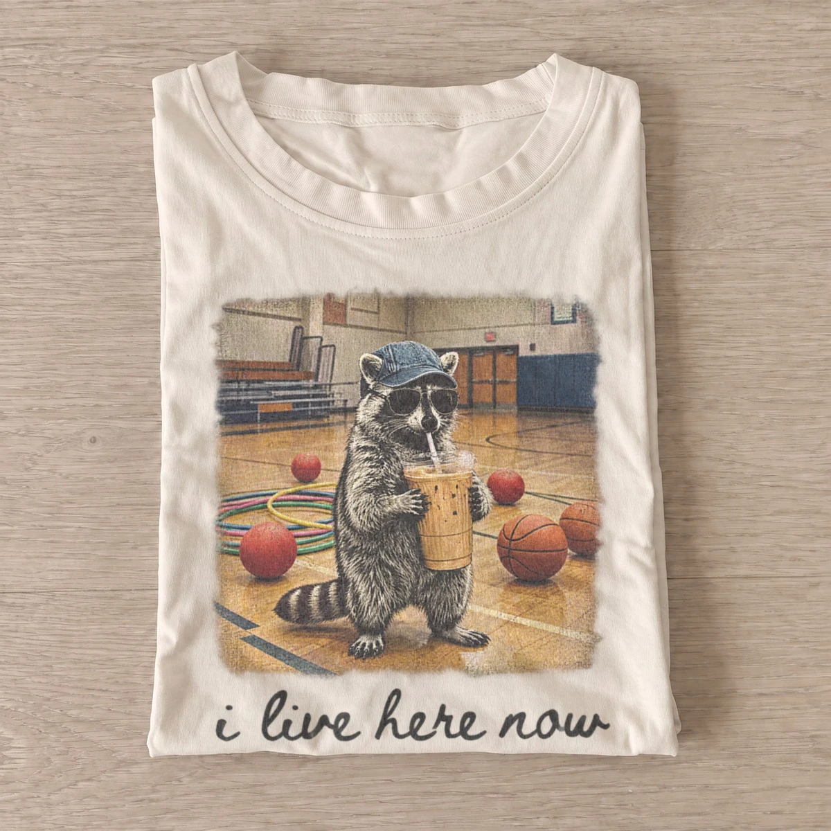 I live here now iced coffee raccoon t shirt sand20iced coffee raccoon.webp